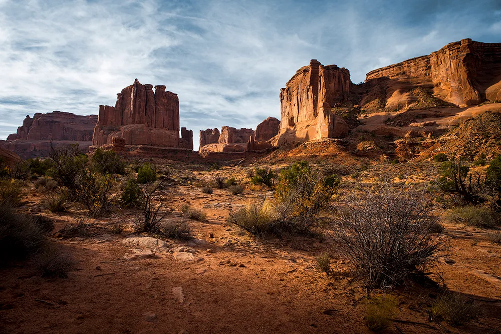 Red sandstone monoliths rising above high desert scrub, lit low and warm beneath a streaked blue sky