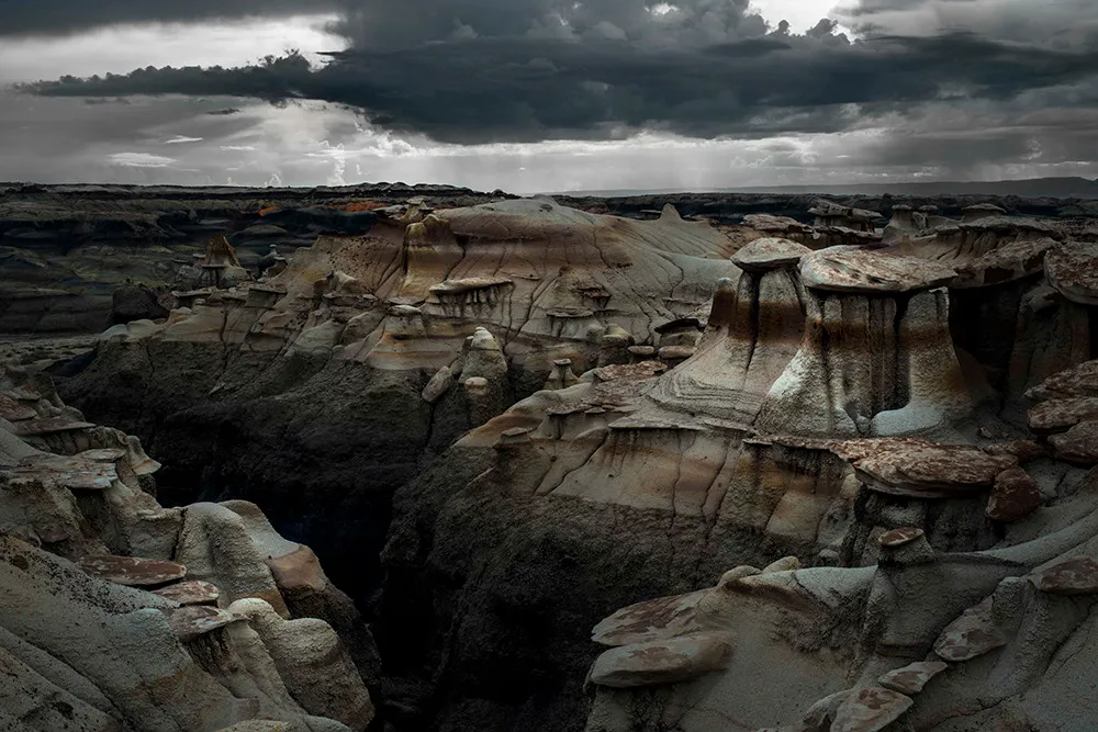 Grey and ochre badlands hoodoos beneath a heavy storm sky, with rain falling on the far horizon