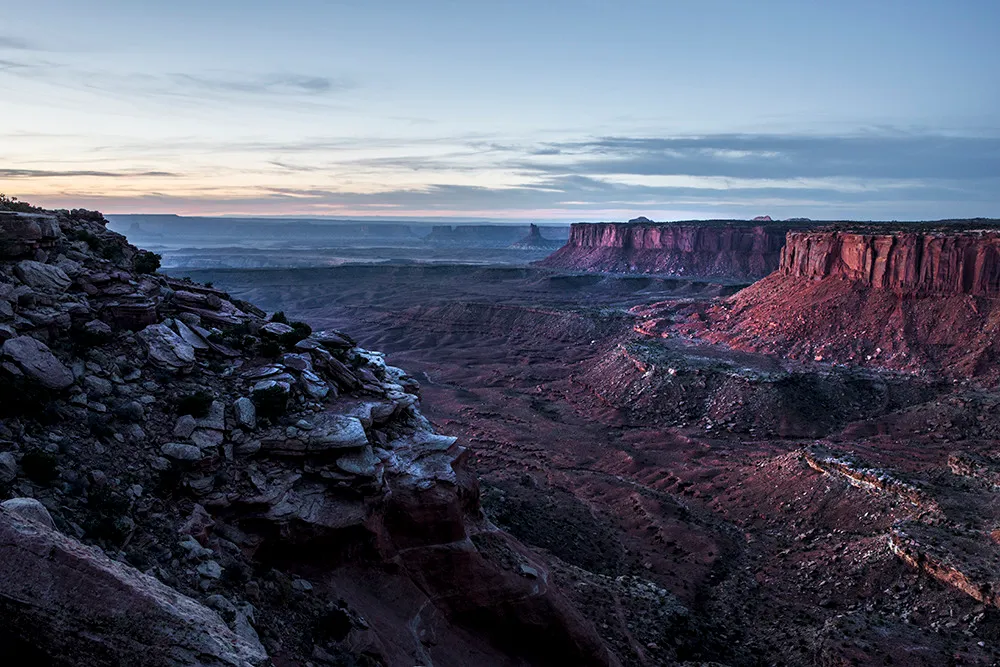 Layered mesas and canyon rims receding into blue evening haze, the nearest cliff still catching pink light