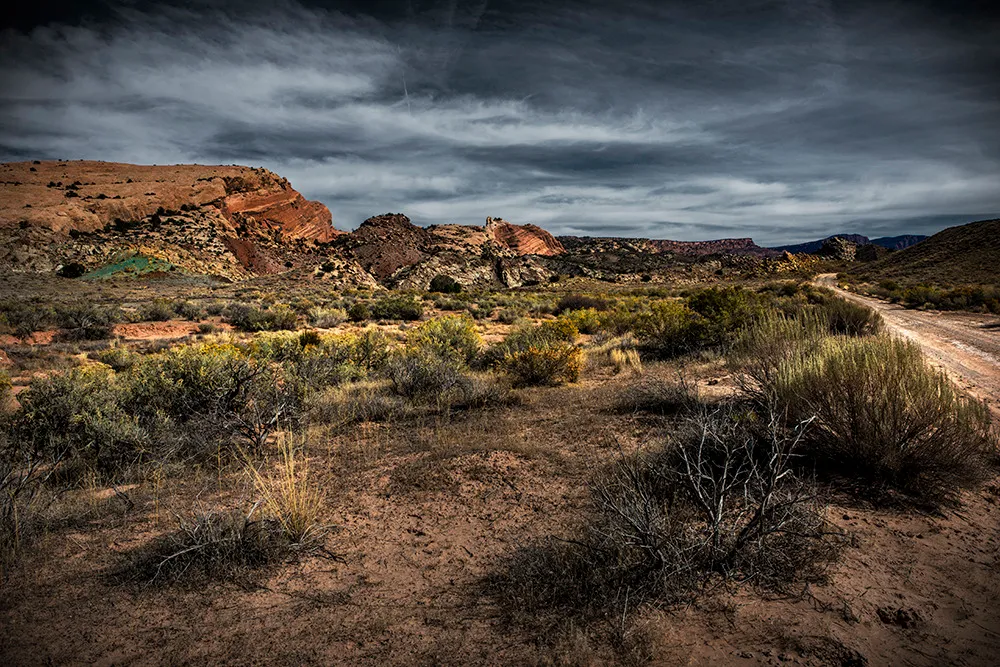 A dirt road running along sagebrush flats toward red rock ridges beneath a bruised, heavy sky