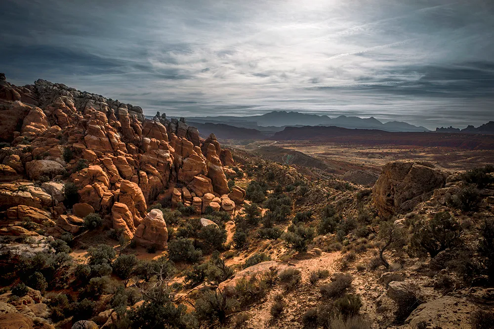 Sunlit sandstone fins in the foreground giving way to a broad valley and snow-touched mountains on the horizon