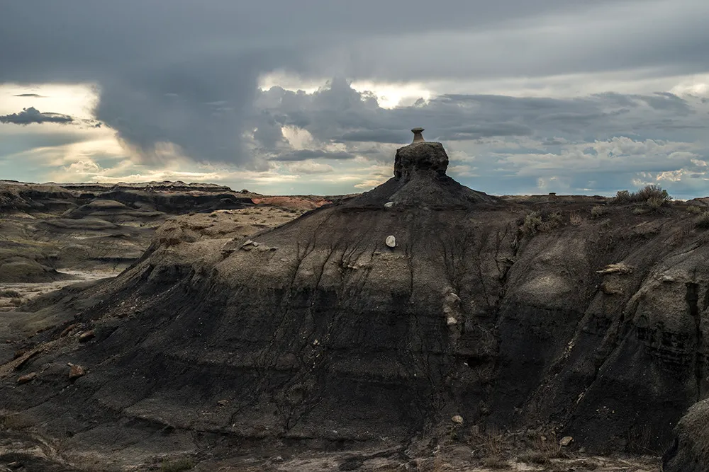 A single capped hoodoo standing on a dark, banded badlands ridge beneath breaking storm clouds
