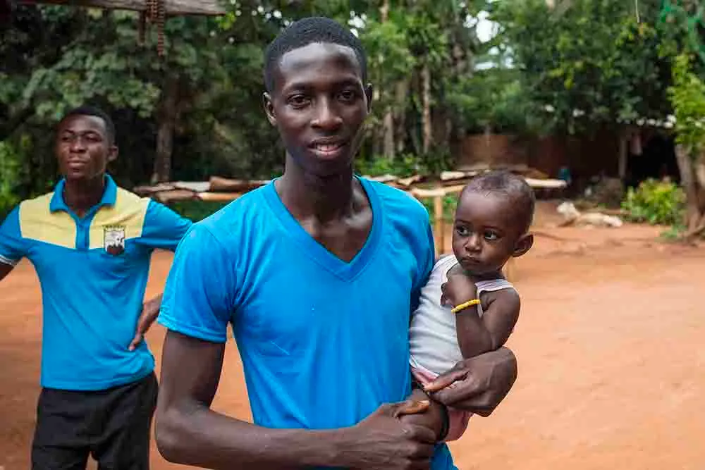 A young man in a blue shirt holding a small child on his hip on a red dirt road, a second young man standing behind them