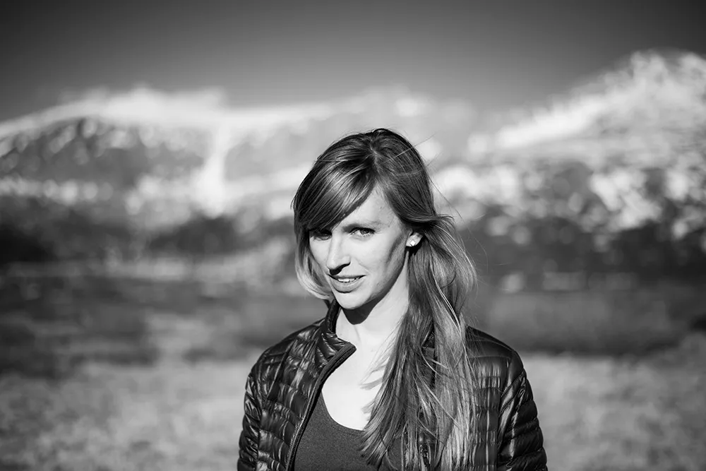 A black and white portrait of a woman in a puffer jacket turning toward the camera, snow-covered peaks blurred behind her