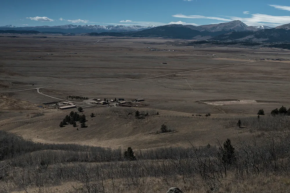 A small ranch and its outbuildings scattered across a broad brown high-country valley below a snow-covered range