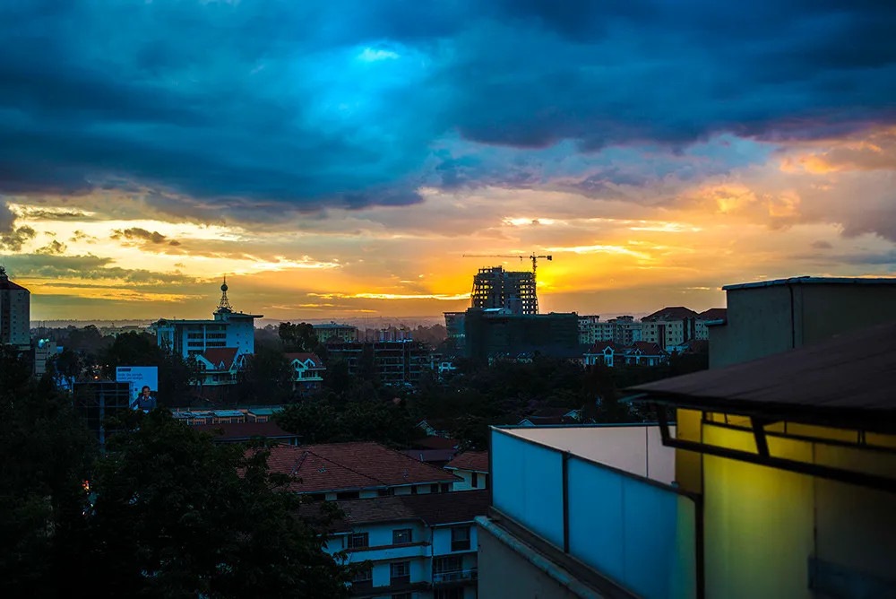 A city skyline of low buildings and a construction crane silhouetted against a gold and blue sunset, seen from a rooftop