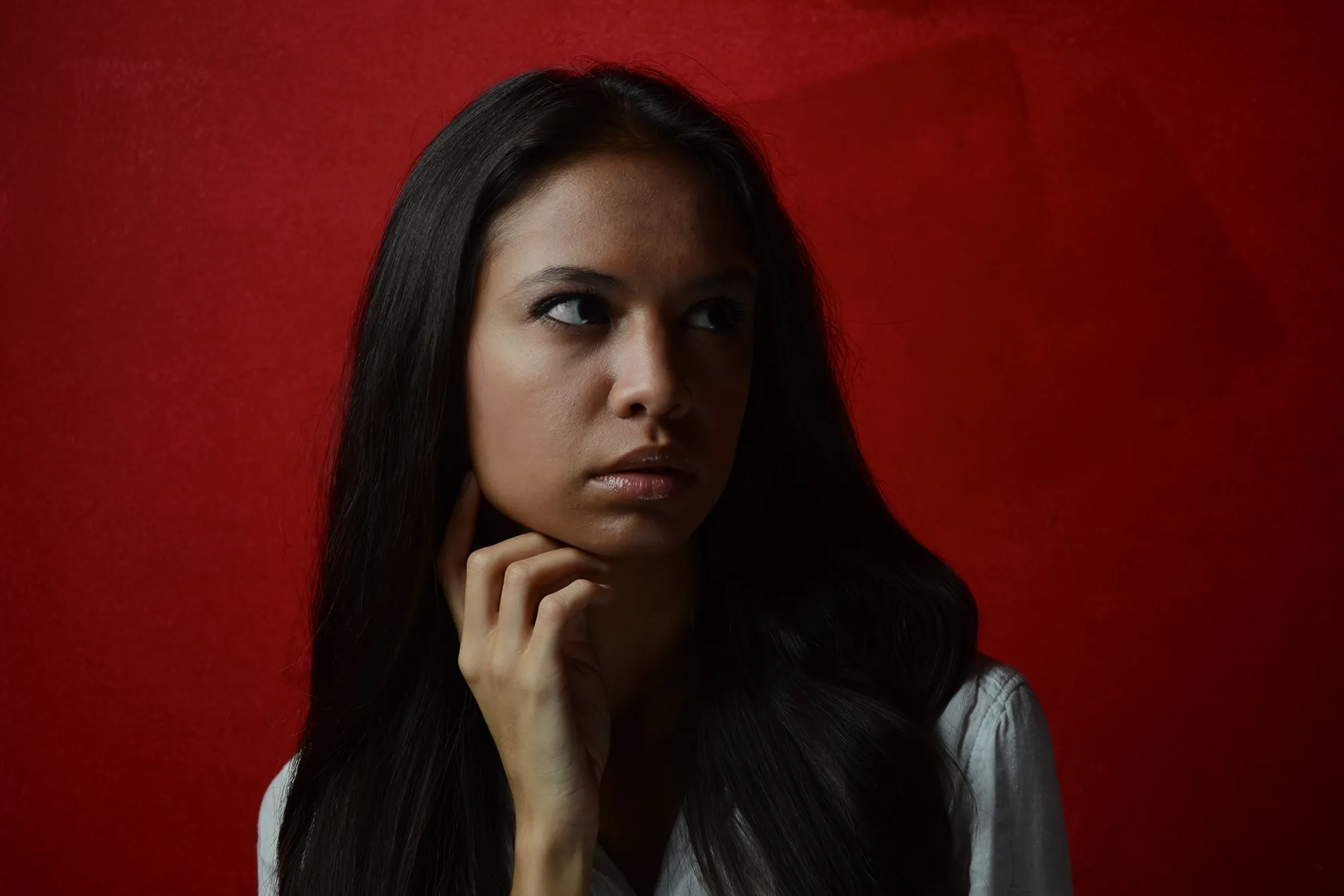 A woman with long dark hair rests her chin on her hand and looks off-camera against a deep red backdrop