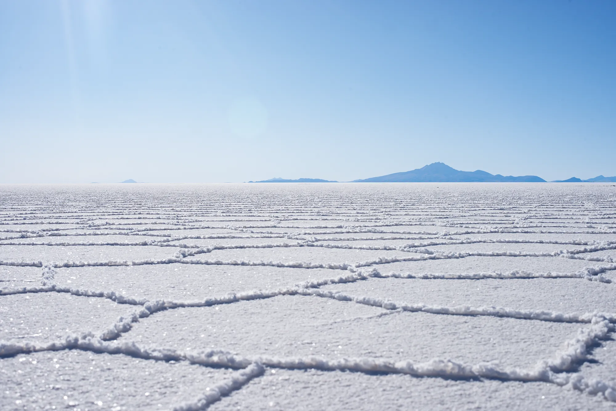 Hexagonal ridges patterning the crust of a vast white salt flat, with distant blue mountains along the horizon