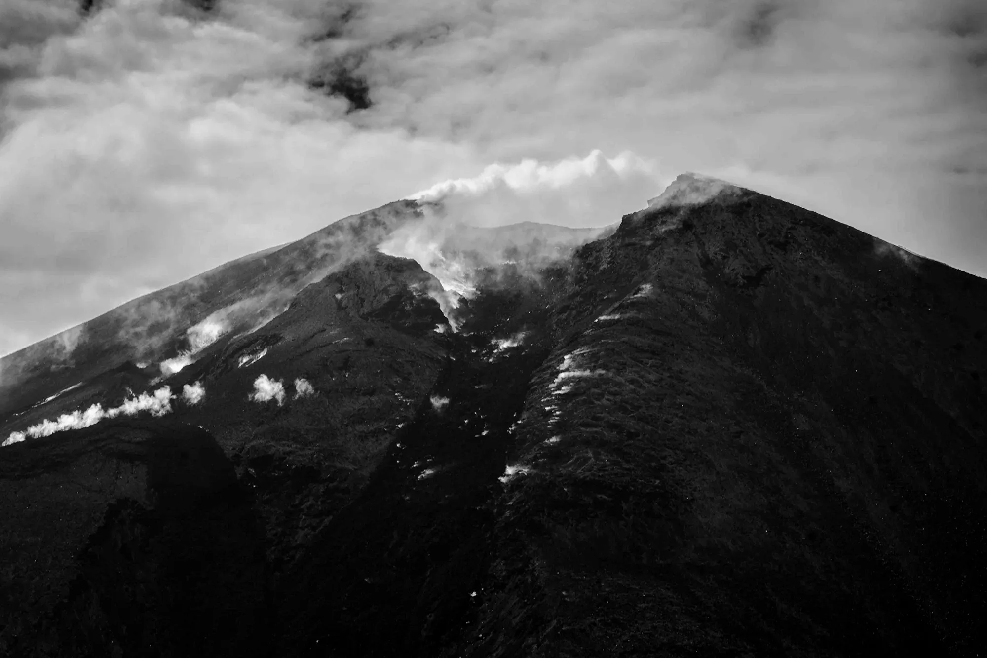 Steam venting from fissures along the dark summit ridge of a volcano, rendered in black and white